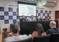 Três homens usando ternos sentados em uma bancada e uma mulher à direita; na plateia, há três pessoas na primeira fila; atrás dos homens da bancada, há uma grande tela de projeção