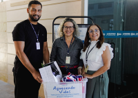 Três pessoas sorridentes (duas mulheres e um homem) posam ao lado de uma caixa transparente com roupas e mantas. Na caixa, há o nome da campanha: “Aquecendo Vidas – Todos juntos para aquecer o inverno de quem mais precisa.” Ao fundo, vê-se parte da estrutura de um prédio institucional.