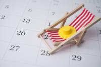 Imagem de uma cadeira de praia de madeira com assento listrado em vermelho e branca e um chapéu amarelo em cima dessa cadeira; a cadeira está sobre um calendário branco
