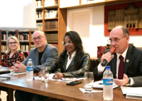 A imagem mostra quatro pessoas sentadas lado a lado em uma mesa, participando de um evento ou reunião. Elas estão em um ambiente com prateleiras cheias de livros ao fundo. Todas estão sorrindo ou com expressão atenta. Uma das pessoas (à direita) está falando ao microfone. Há garrafas de água, copos e papéis sobre a mesa.