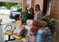 Em destaque, três volumes de alimentos, com fuba ao centro, feijão à direita e arroz à esquerda; atrás dos alimentos, três mulheres morenas abraçadas