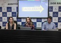Foto de três pessoas sentadas à mesa, sendo duas mulheres à esquerda e um home à direita. Atrás deles, um projetor com o nome acelera 19 