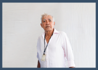 Homem de pele clara e cabelos grisalhos aparece em pé diante de um fundo branco. Ele veste uma camisa social branca, levemente aberta no colarinho, e usa um cordão com crachá no pescoço. Seu semblante é sério e ele olha ligeiramente para cima.
