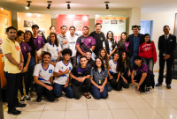 Grupo de estudantes e adultos posam sorrindo para foto em frente a painéis coloridos com conteúdo histórico em ambiente interno bem iluminado.