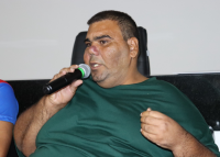 Foto de um homem morendo, cabelos pretos, gordo, sentado de camisa verde e seguraando um microfone preto na mão direita.