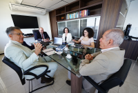A foto mostra cinco pessoas reunidas em torno de uma mesa de vidro em um ambiente corporativo. Duas delas, em primeiro plano, vestem ternos claros e parecem estar em diálogo. Os outros três participantes, ao fundo, estão atentos e sorrindo, com notebooks e anotações sobre a mesa. O clima é de reunião formal, porém descontraída.