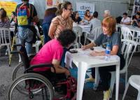 A foto mostra um atendimento voltado à inclusão de pessoas com deficiência em um evento de empregabilidade. Em primeiro plano, uma mulher em cadeira de rodas, vestindo camiseta rosa, conversa com uma atendente sorridente, que escreve em uma ficha sobre a mesa branca. Ao redor, outras pessoas participam do evento, algumas em cadeiras de rodas ou com muletas, reforçando o caráter inclusivo da iniciativa. No fundo, banners de empresas e programas de inclusão são visíveis.