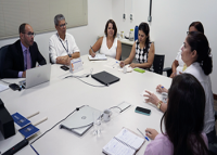 Foto de várias pessoas sentadas ao redor de uma mesa, sendo dois homens e cinco mulheres. Só há um homem de terno e restante das pessoas usa roupas informais