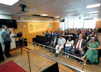 Foto de um auditório com público atento assistindo a uma palestra. À esquerda, um homem de blazer fala ao microfone. À direita, pessoas sentadas, entre elas um homem de terno e uma mulher de vestido verde. Ao fundo, uma janela grande e um painel com o nome do auditória Des. José Abílio Neves Souza. O ambiente é claro e bem organizado.