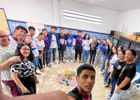 A imagem mostra um grupo de pessoas, entre jovens e adultos, formando um círculo em uma sala de aula. Todos estão sorrindo e abraçados, olhando para a câmera, que está sendo segurada por um dos jovens em primeiro plano. No centro do círculo, há um arranjo colorido no chão, com flores, objetos decorativos e cartões dispostos em círculos, formando uma mandala. O ambiente é claro, com paredes azuis e armários com livros ao fundo. A imagem transmite união e acolhimento.