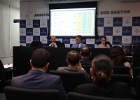 A imagem mostra um auditório onde ocorre uma apresentação institucional. Quatro pessoas estão sentadas à mesa diretiva, sendo dois homens ao centro — um deles falando ao microfone — e uma mulher à direita operando um notebook. Ao fundo, uma projeção exibe um slide com texto e blocos coloridos, indicando a exposição de um projeto ou plano. O público, composto por homens e mulheres em trajes formais, acompanha atentamente a apresentação. A parede ao fundo exibe o nome “Auditório” e parte da identidade visual 