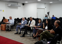 A imagem mostra uma sala de reuniões com cadeiras organizadas em fileiras. No primeiro plano, sete pessoas estão sentadas em cadeiras, formando uma espécie de semicírculo. Algumas delas seguram pastas e blocos de anotações. Uma mulher, à direita, segura um microfone e fala, enquanto os demais parecem ouvir com atenção. Ao fundo, há mais cadeiras vazias, duas pessoas sentadas próximas a uma câmera em tripé e, na parede, um aparelho de ar-condicionado. O ambiente é iluminado por luzes de teto e tem piso de ma