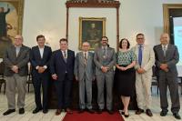  imagem mostra oito pessoas em pé, lado a lado, posando para foto em um ambiente formal. Todos estão vestidos com roupas sociais; a maioria usa terno e gravata, e uma das pessoas veste blusa verde com estampa de bolinhas e saia preta. Três homens usam no pescoço medalhas douradas com fita azul, indicando uma solenidade. Ao fundo, há quadros de figuras históricas e uma parede clara. O piso é de ladrilhos brancos, e um tapete vermelho cobre parte do chão em frente ao grupo.