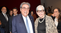Um homem e uma mulher posam sorrindo para a foto em um ambiente formal, provavelmente um evento. O homem veste terno azul-marinho, camisa branca e gravata azul-clara; a mulher tem cabelo branco curto, usa óculos de armação grossa preta, colar de pedras e um casaco estampado em preto e bege sobre blusa preta. Outras pessoas aparecem desfocadas ao fundo.