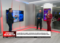 rês apresentadores estão em um estúdio de televisão com iluminação clara e detalhes em vermelho. Eles sorriem e conversam enquanto uma tela exibe a imagem da campanha "Guardiões do Saber", que arrecada kits escolares para crianças em situação de vulnerabilidade. No rodapé da tela, há a tarja do programa Boletim Gazeta com a manchete da campanha e um pequeno noticiário rolando. Ao fundo, vê-se parte da redação do jornal.