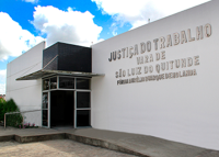 Fachada de um prédio com paredes brancas, identificado como sede da Justiça do Trabalho. Na parede está escrito: "Justiça do Trabalho – Vara de São Luiz do Quitunde – Fórum Aurélio Buarque de Holanda". O céu está azul com nuvens brancas. A entrada principal tem uma cobertura metálica e portas de vidro.