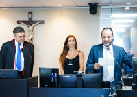 Homem alto de paletó preto, blusa bracan e gravata azul à esquerda; ao centro, mulher baixa de vestido preto e cabelos ruivos, à direita, homem moreno de blazer azul e camisa branca. Todos estão em pé.