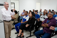 Homem idosos magro, de camisa branca e calça cáqui, em pé minsitrando palestra para homens e mulheres sentados.