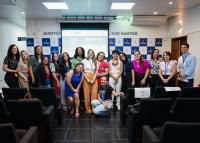 Na imagem, um grupo de pessoas está reunido em um auditório, posando para a foto em frente a um telão e a um painel com o logotipo “Rede Estadual”. Elas estão sorrindo e aparentam estar em um momento de confraternização após uma atividade ou reunião. Há homens e mulheres, a maioria em pé, e uma pessoa agachada à frente. O ambiente é iluminado, com cadeiras pretas no primeiro plano.