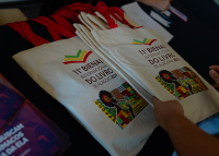 A imagem mostra algumas ecobags (sacolas de tecido reutilizáveis) dispostas sobre uma mesa. As bolsas têm alças vermelhas e estampa colorida na parte frontal, com elementos gráficos que remetem à cultura afro-brasileira. No centro, está o logotipo e o texto: “11ª Bienal Internacional do Livro de Alagoas 2023”.  À esquerda, parcialmente visíveis, estão alguns materiais gráficos ou panfletos, enquanto à direita aparece a mão de uma pessoa segurando uma das sacolas. 
