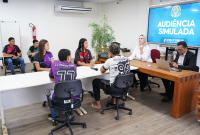 Estudantes participam de uma audiência simulada em uma sala de audiência. Dois magistrados estão à mesa conduzindo a atividade, enquanto os jovens, sentados em cadeiras, representam as partes do processo. No telão ao fundo, aparece o texto 'Audiência Simulada – Justiça do Trabalho'."
