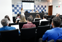 A foto mostra um auditório durante uma reunião ou palestra. No ambiente, pessoas estão sentadas em poltronas pretas, voltadas para a frente, onde há uma mesa com três pessoas posicionadas — duas sentadas e uma em pé, falando ao microfone.  No fundo, um telão exibe uma projeção com informações digitais, indicando que a apresentação conta com apoio de recursos audiovisuais. O auditório tem paredes claras, com o logotipo do TRT da 19ª Região em um painel azul e branco, e a identificação “Auditório … dos Santos