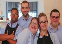 A foto mostra cinco pessoas, todas sorridentes e reunidas em um ambiente interno, que parece ser um restaurante. Elas usam camisas sociais azuis claras, e duas delas também vestem aventais pretos, sugerindo que trabalham no local. O grupo transmite alegria, acolhimento e companheirismo, reforçando um clima de inclusão e diversidade. Ao fundo, é possível ver janelas grandes que deixam entrar bastante luz natural.