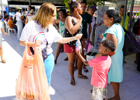 A foto mostra uma cena de solidariedade em um espaço público. Uma mulher, usando óculos escuros e camiseta branca estampada, segura uma sacola grande em uma das mãos e entrega uma garrafa de água a um menino pequeno, que estende o braço para recebê-la. Ao fundo, há várias pessoas, incluindo mulheres e crianças, algumas em pé e outras sentadas, em um ambiente movimentado que parece ser uma ação social ou comunitária. O clima transmite cuidado, acolhimento e apoio às pessoas presentes.