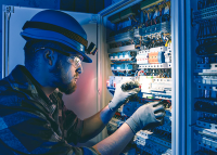 Há um homem. Ele é um eletricista profissional e está concentrado em seu trabalho. Eleusa um capacete de segurança e luvas de proteção isolantes para evitar choques. Com as mãos, ele mexe com cuidado nos fios e disjuntores dentro de um grande quadro de energia elétrica metálico, garantindo o funcionamento seguro da energia