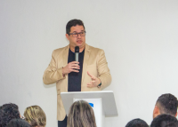 Homem alto de cabelo preto em pé proferindo palestra com um microfone na mão direita e vestindo blusa preta e blazer creme