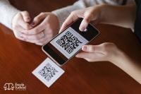Imagem mostra uma pessoa usando um celular para escanear um QR Code impresso em um cartão branco, sobre uma mesa de madeira. No canto inferior esquerdo, aparece o logotipo da Escola Judicial do TRT-AL. Outra pessoa está com as mãos apoiadas sobre a mesa, observando a ação.