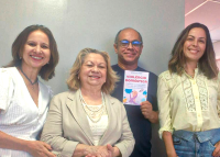 A imagem mostra quatro pessoas sorrindo para a câmera. Três mulheres e um homem, que segura uma cartilha com o título **“Violência Doméstica”**.