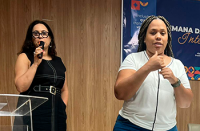 Mulher de vestido preto e cabelo preto com microfone na mão e uma mulher negra fazendo libras