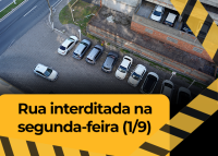 Rua com carros estacionados. Há uma tarja amarela escrito "Rua interditada na segunda-feira (1/9), com letras pretas.