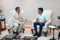 Dois homens estão sentados frente a frente em poltronas claras, em uma sala de reunião. À esquerda, um homem de terno bege com gravata laranja segura papéis e conversa. À direita, um homem de camisa azul clara de mangas dobradas e calça jeans ouve atentamente. Entre eles há uma mesinha com caderno e canetas.