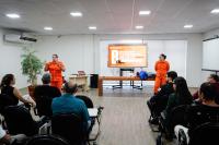 Duas mulheres de macacão laranja ministrando aula para uma plateia formada por homens e mulheres, sendo uma do lado direito e a outra do lada esquerdo