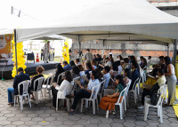 Homem de terno cinza claro em cima do palco discursando com o microfone na mão direita; à frente dele, muitos homens e mulheres sentados