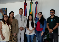 Na foto, um homem alto de terno cinza ao meio de duas mulheres ao seu lado direito e duas ao lado esquerdo; no extremo direito, um homem moreno de cabelos pretos e blusa verde, atrás deles, três bandeiras coloridas de verde, amarelo, vermelho, azul e branco