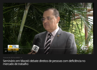 Há um homem de terno cinza, camisa branca e gravata listrada em preto e branco, sendo entrevistado por uma repórter que segura um microfone da TV Globo. Ele está em frente a plantas verdes, em um ambiente externo. Na parte inferior da tela aparece a legenda: “Seminário debate direitos de pessoas com deficiência no mercado de trabalho”.