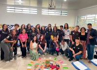 Há um grupo, a maioria jovens estudantes, posando sorridentes em uma sala de aula. Eles estão reunidos em pé e sentados, alguns vestindo uniformes escolares pretos e outros roupas casuais. No centro do chão, há uma roda decorada com tecidos coloridos, papéis e objetos simbólicos, formando um círculo. O ambiente é claro, com paredes azulejadas em azul e amarelo e janelas grandes que deixam entrar bastante luz natural.