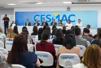Há um saão repleto de pessoas sentadas. Há uma mesa em destque com várias pessoas.