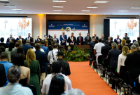 À frente, várias autoridades em pé atrás de uma mesa de honra; à frente das autoridades e de costas para a foto, várias pessoas entre homens e mulheres