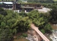 Fotografia aérea da Usina Santa Clotilde, em Alagoas. A imagem mostra parte da estrutura industrial ao fundo, cercada por vegetação verde. Em primeiro plano, há um rio e uma pequena ponte de concreto ligando as margens.