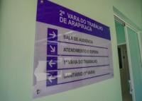 A imagem mostra uma placa de sinalização interna de um prédio do Tribunal Regional do Trabalho. Na parte superior, em fundo roxo, está escrito:  “2ª VARA DO TRABALHO DE ARAPIRACA”.  Abaixo, em fundo branco, há setas indicando a direção de diferentes setores:  Sala de Audiência (seta para a direita)  Atendimento e SEPEJA (seta para a direita)  1ª Vara do Trabalho (seta para a esquerda)  Sanitário (1ª Vara) (seta para a esquerda)  A placa está fixada em uma parede de cor verde-clara.i