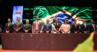 Homens e mulheres de pé em posse solene em frente a um telão com a imagem da bandeira do Brasil