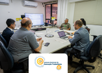 Foto de quatro homens e uma mulher todos sentado de costas em uma mesa de sala de audiência; à frente deles, um monitor.