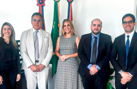 Na imagem, três homens de terno, sendo dois de ternos pretos e um de terno cinza claro, além de duas mulheres loiras, uma de vestido cinza estampado e uma usando terno preto