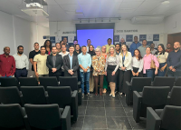 Há um grupo de pessoas posando para foto em um auditório. Elas estão organizadas em duas fileiras, com algumas pessoas em pé atrás das cadeiras e outras à frente delas. No centro, há três mulheres e um homem em destaque, com roupas sociais. Ao fundo, vê-se uma tela de projeção azul. O ambiente é iluminado e transmite a ideia de um encontro formal ou institucional.