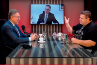 A imagem mostra um estúdio de gravação de podcast, com duas pessoas sentadas frente a frente em uma mesa de vidro, equipada com microfones e copos de água.  À esquerda, um homem de terno azul sorri enquanto fala. À direita, outro homem, de camiseta preta, gesticula com a mão levantada, participando ativamente da conversa.  No centro da cena, atrás da mesa, há uma televisão fixada na parede exibindo um terceiro homem, também de terno, que aparece em chamada de vídeo, completando o bate-papo.  