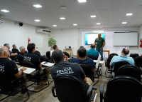 Há uma sala de aula com várias pessoas sentadas. Elas assitem uma pessoa falando que está  de pé à frente delas.